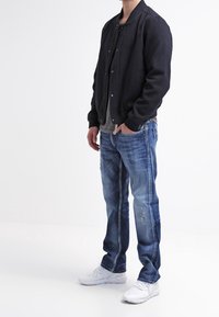 Svart bomberjacka med knappar, grå skjorta under, blå denimjeans med slitna detaljer och vita sneakers. Avslappnad outfit totalt sett.