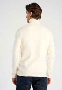 Cremefarbener Rollkragenpullover, Strickstruktur, figurbetontes Design, gerippte Bündchen und Saum, von hinten mit sichtbarer dunkler Hose.