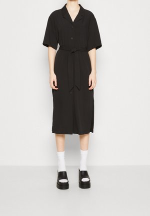 Zwart overhemdjurk met korte mouwen, zelfstrikceintuur, knielengte, zijsplitten, gecombineerd met zwarte sleehakken en witte sokken.