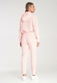 Jasnoróżowy dres wykonany z miękkiego materiału, składający się z bluzy z kapturem i dopasowanych spodni z rozcięciami przy kostkach, w połączeniu z białymi butami.