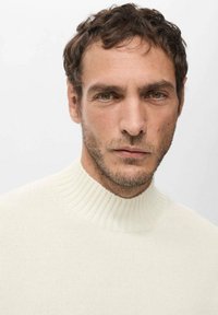 Suéter de punto acanalado color crema con cuello alto. Textura suave, diseño ajustado, costuras mínimas y brillo sutil. La modelo tiene el cabello corto.