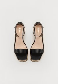 Sandalias de cuña de cuero negro con una correa al tobillo, que presentan una banda frontal ancha y una suela de yute texturizada. En el interior se muestra el logotipo en dorado.