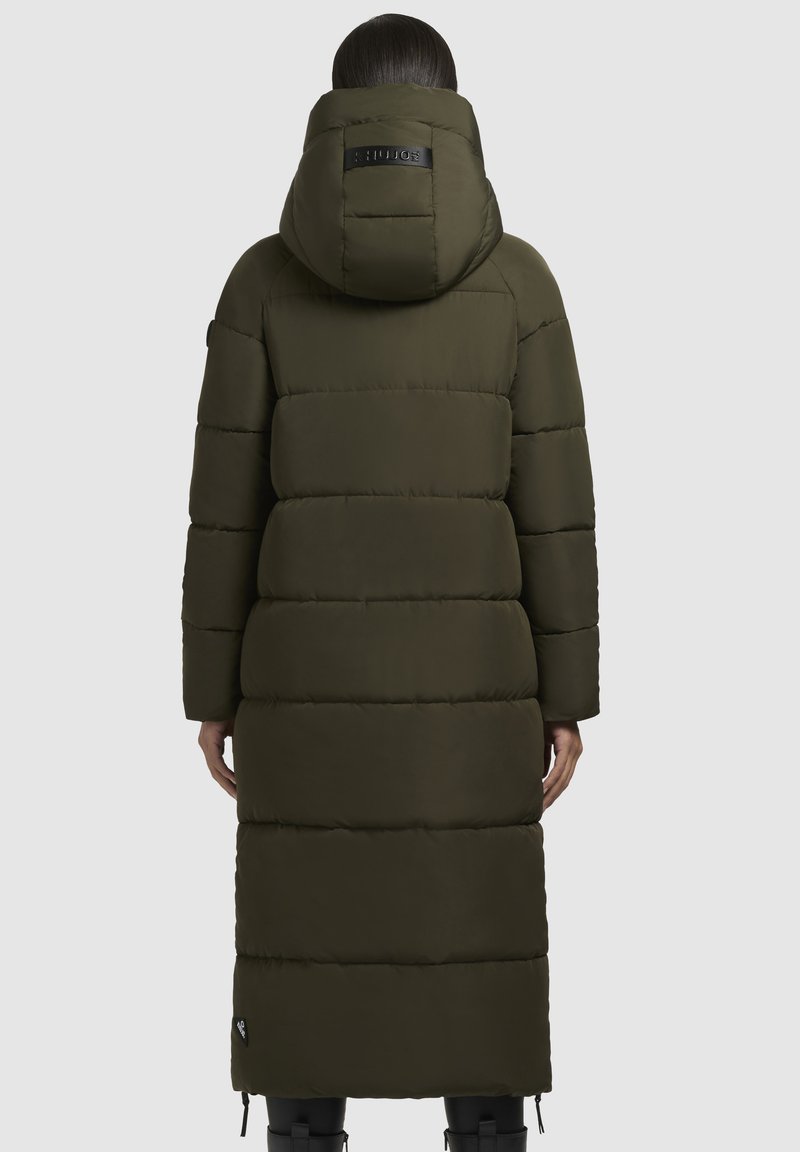Person, der einen langen, dunkelgrünen, gepolsterten Wintermantel mit Kapuze trägt, von hinten vor einem schlichten, hellen Hintergrund gezeigt.