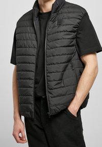 Černá péřová vesta s horizontálně prošívaným vzorem, vysokým límcem a postranními zipovými kapsami, nošená přes černé triko. Hladký, lehký materiál.