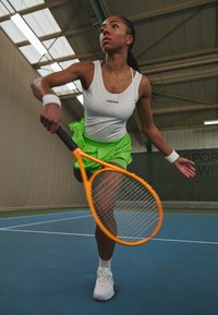 Athlète portant un débardeur blanc et une jupe vert fluo, tenant une raquette de tennis orange, sur un court intérieur bleu avec une surface lisse.
