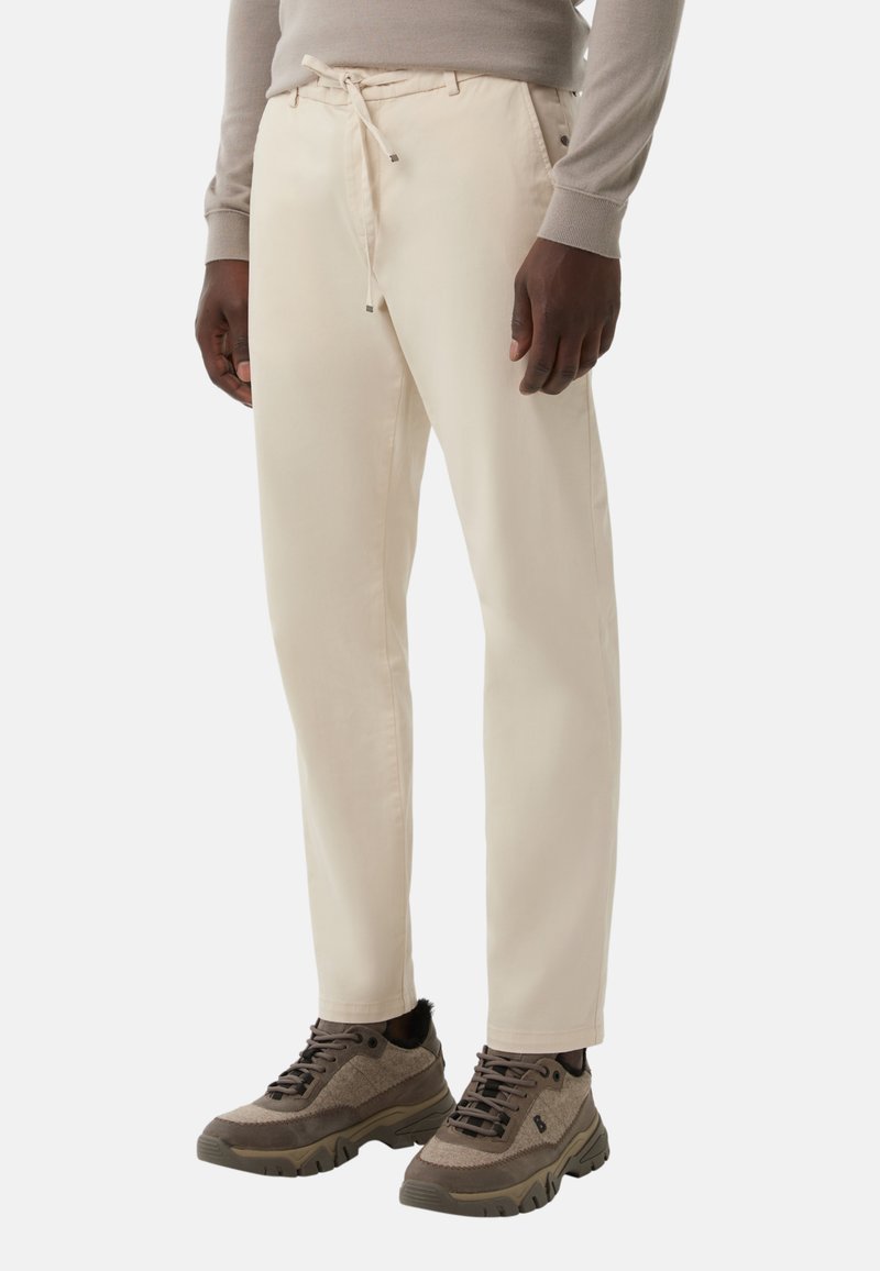 Homme portant un pantalon beige à cordon de serrage et des baskets taupe à semelles épaisses, se tenant devant un fond blanc uni.