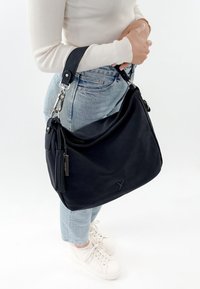 Kobieta w jasnych dżinsach, białych sneakersach i kremowym swetrze trzyma dużą czarną torbę na ramię z frędzlami i srebrnymi detalami.