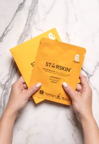 Felgeel bio-cellulose gezichtsmasker verpakking met tekst. Twee zakjes vastgehouden tussen handen tegen een witte marmeren ondergrond.
