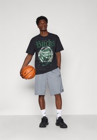 Sort T-shirt med grøn "Bucks" grafik, grå sportsshorts, sorte sneakers, mens han holder en basketball. Simpelt design uden synlige mønstre.