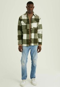 Groen en wit geruit flanellen shirt, lange mouwen, voorzakken, gecombineerd met een tan ribbeltrui en versleten lichtblauwe jeans.