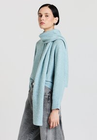 Hellblauer Pullover mit einem dicken umgeschlagenen Kragen, langen Ärmeln und einem drapierten Schal-Detail; kombiniert mit hoch geschnittenen grauen Jeans.