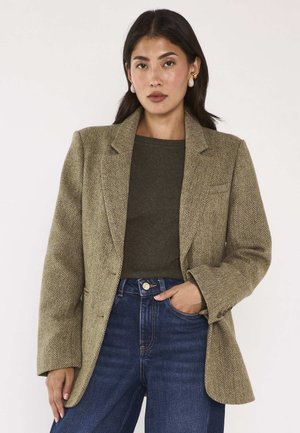 Jonge vrouw met donker haar draagt gouden oorbellen, een olijfgroene trui, een beige blazer met patroon en blauwe high-waist jeans, poseert met één hand in de zak.