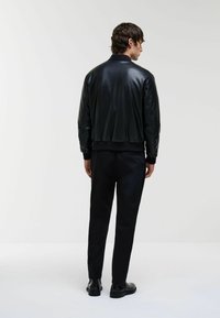 Jachetă bomber neagră din piele fină, cu manșete și tiv ribbed, având un design minimalist și o siluetă ajustată. Purtată cu pantaloni închiși la culoare.