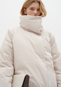 Šviesiai rožinė paminkštinta striukė su aukštu apykakle, kuri turi kupolo formos sagas ir glotnų, blizgantį paviršių. Modelis laiko dvišalį rankinę.