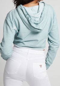 Felpa cropped azzurra chiara con una texture morbida e cappuccio con cordino, indossata su jeans bianchi con tasche posteriori e un piccolo logo.