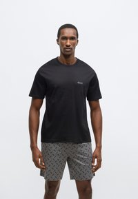 Čovjek odjeven u crnu BOSS majicu i sive kratke hlače s uzorkom crnog BOSS logotipa, stoji ispred jednostavne bijele pozadine.