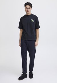 Fekete pamut póló rövid ujjakkal és kerek nyakkivágással, zöld szív logóval. Tengerészkék slim-fit nadrággal és fekete cipővel párosítva.