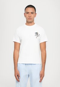 White cotton t-shirt with short sleeves and round neck. Features a colorful embroidered design on the left chest. Model wears striped pajama pants.