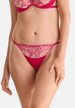 Femme portant un ensemble de lingerie en dentelle rose, montrant la taille et le haut des cuisses sur un fond uni clair.