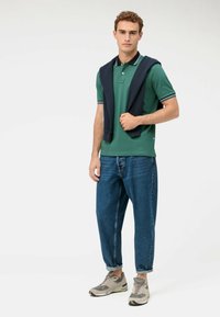 Polo verde con colletto a righe bianche e blu marina, abbinato a jeans blu larghi e sneakers grigie. Maglione poggiato sulle spalle.
