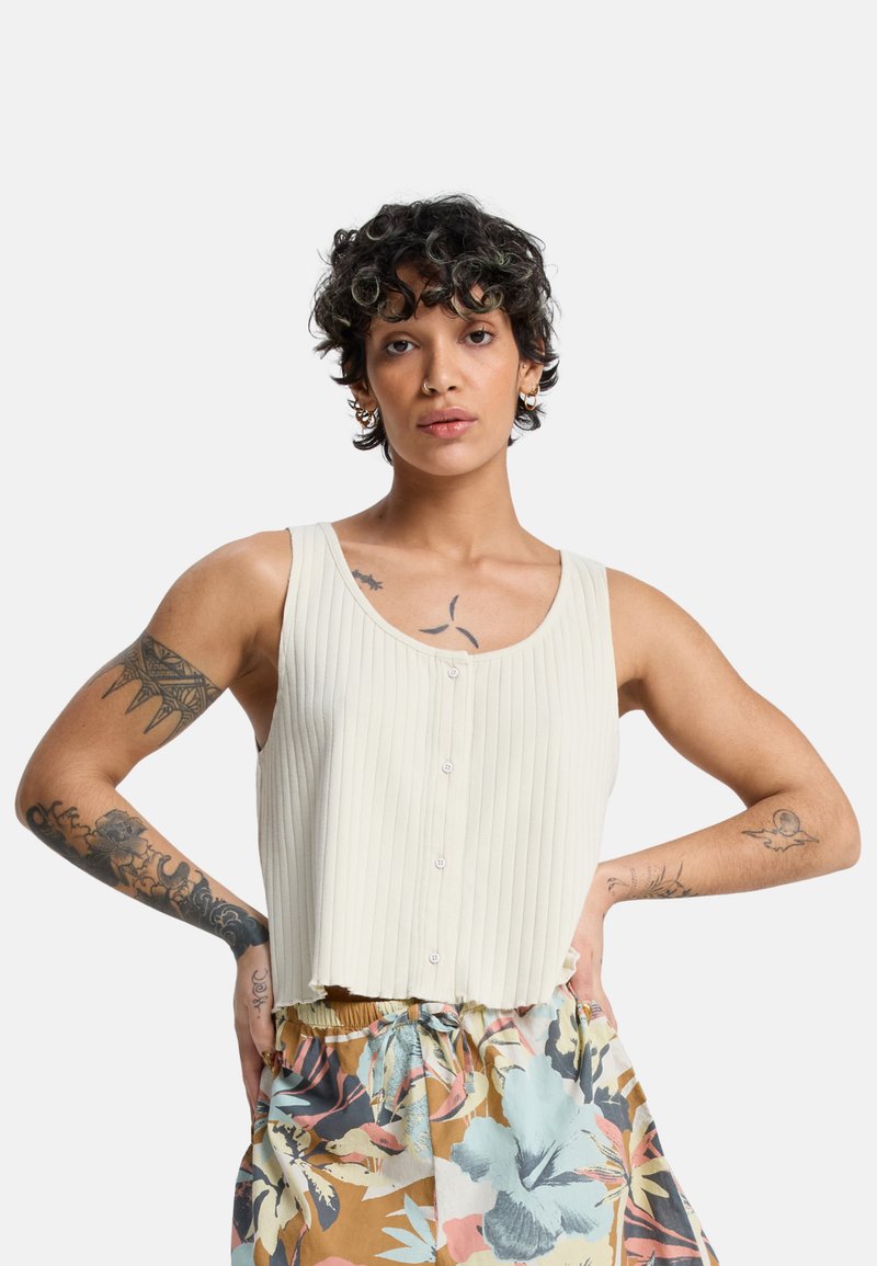 Άτομο με κοντά κατσαρά μαλλιά, φορώντας ένα αμάνικο ριμπερέ εκρού τοπ και παντελόνια με λουλουδάτο σχέδιο, στέκεται με τα χέρια στη μέση.