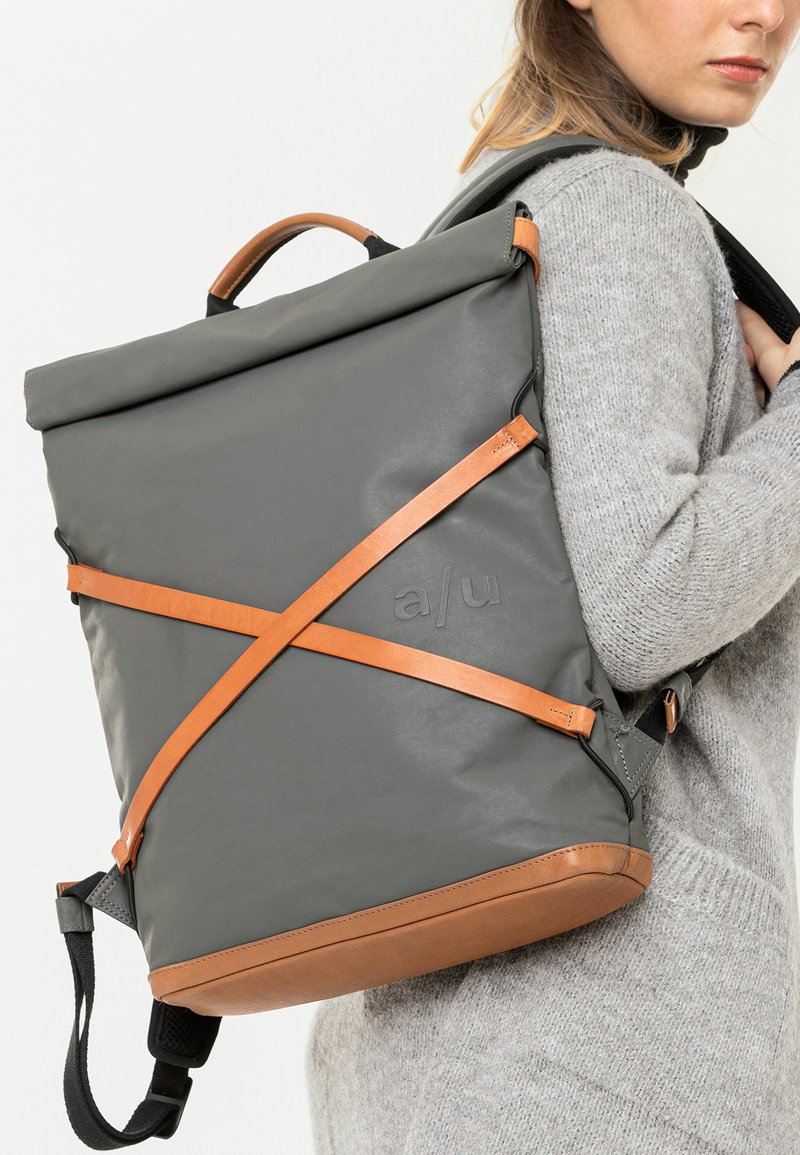 Grau roll-top Rucksack mit braunen Lederakzenten und Crossbody-Trägern. Verfügt über eine glatte Textur und gepolsterte Schulterriemen.