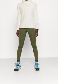 Cremefarbener Fleece-Turtleneck, olivgrüne Leggings und blaue gemusterte Sportschuhe. Strukturierten Oberfläche oben, eng anliegende Hosen mit Nahtdetails.