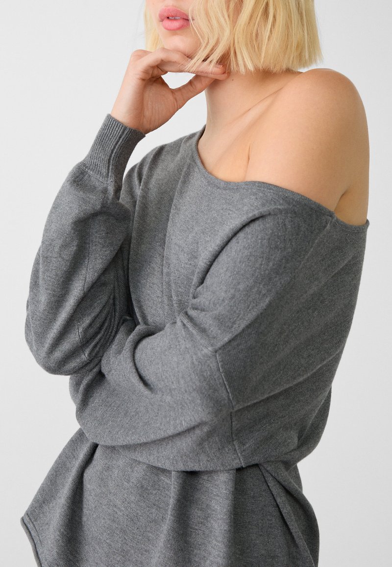 Szary sweter bez ramion z długimi rękawami, prążkowanymi mankietami i luźnym krojem. Materiał wydaje się miękki i teksturowany, z delikatnym spadkiem.