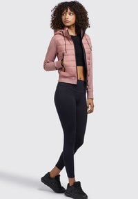 Pinker gepolsterter Reißverschlussjacke mit Kapuze, schwarzem Crop-Top und schwarzen Leggings. Schwarze Sportschuhe runden das Outfit ab.
