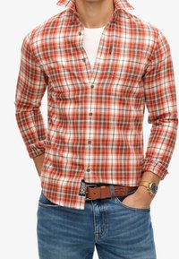 Camicia a quadri rossa e bianca con accenti azzurri, design con bottoni, maniche lunghe e tasca sul petto. Indossata con una cintura marrone e un orologio.