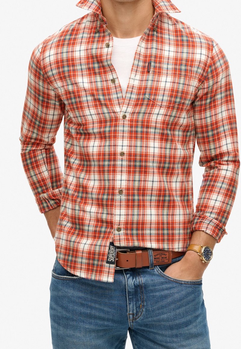 Camicia a quadri rossa e bianca con accenti azzurri, design con bottoni, maniche lunghe e tasca sul petto. Indossata con una cintura marrone e un orologio.