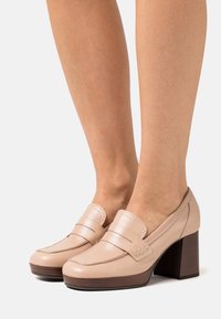 Beige leather loafers with a rounded toe, detailed stitching, and a brown block heel. Textured finish enhances visual interest.