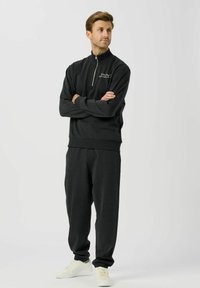 Temno siva trenirka iz mehke tkanine z zadrgo na ovratniku, širokimi hlačami in belimi supergami. Minimalističen dizajn.