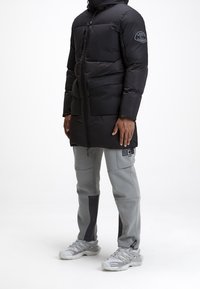 Manteau matelassé noir avec capuche, fermeture zippée, deux poches avant et écusson de marque sur la manche ; assorti à un pantalon de jogging gris et des baskets.