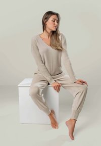 Frau mit langen Haaren, die einen beigen Strickpullover und eine Hose trägt, sitzt barfuß auf einem weißen Würfel und schaut mit geschlossenen Augen seitlich weg.