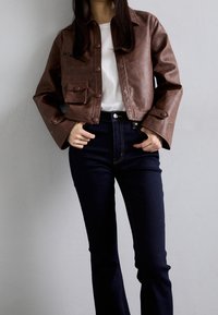 Veste courte en cuir marron avec fermetures à boutons, deux poches avant et poignets évasés, associée à un jean foncé taille haute.