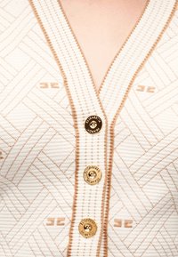 Κοντινή λήψη λευκού υφάσματος με υφή και μπεζ γεωμετρική ραφή, με χρυσά κουμπιά που φέρουν την εγχάρακτη επιγραφή «Elisabetta Franchi.»