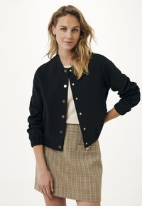 Czarna kurtka bombowiec z złotymi zatrzaskami, ściągaczami u rękawów i skróconym krojem, w połączeniu z teksturowaną, kratkowaną mini spódnicą w beżowych i brązowych odcieniach.