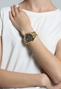 Orologio in metallo dorato con quadrante nero, marcatori in numeri romani e bracciale a maglie, indossato sul polso di un modello contro una maglietta chiara.
