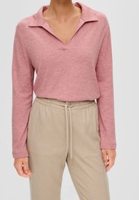 Långärmad, rosa tröja med krage och V-ringning, tillsammans med beige byxor med elastisk midja och snörning. Slät tygtextur.