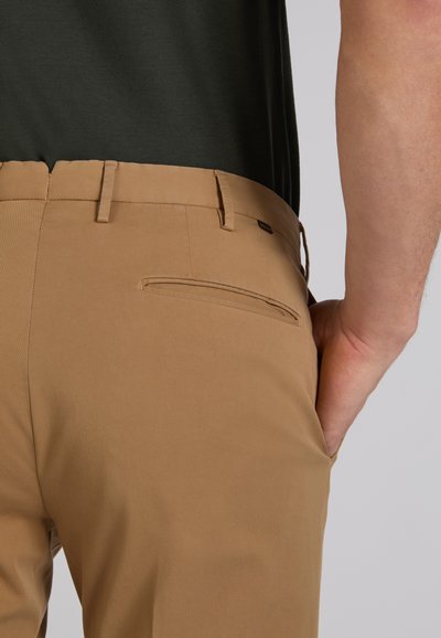 Zbliżenie na osobę w beżowych spodniach z tylną kieszenią z listwą oraz czarnej koszuli, z ręką spoczywającą w przedniej kieszeni, na neutralnym tle.