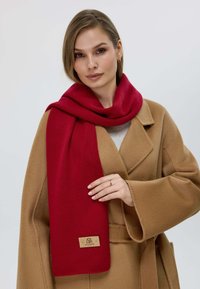 Sciarpa rossa realizzata in morbido materiale, appesa attorno al collo. Presenta una forma rettangolare e un piccolo etichetta con marchio su sfondo beige.
