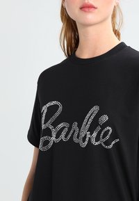 Černé tričko s volným střih, který je zdobený lesklými stříbrnými kamínky ve tvaru kurzívou napsaného slova "Barbie" na přední straně. Krátké rukávy a kulatý výstřih.
