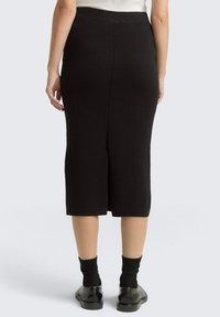 Zwarte gebreide rok tot de knie met een split aan de achterkant, met een strak, gladde textuur en een aangesloten silhouet. Gestyled met zwarte sokken en schoenen.