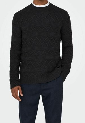 Man draagt een zwarte, getextureerde gebreide trui over een wit ondershirt met donkerblauwe broek, één hand in de zak.