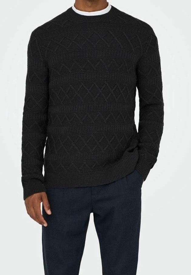 Homme portant un pull en maille noir texturé par-dessus un t-shirt blanc, avec un pantalon bleu foncé, une main dans la poche.