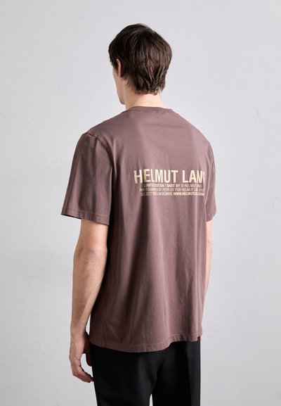 Barna pamut póló, amelynek hátán arany nyomott logó található. Rövid ujjak, laza fazon és kerek nyakkivágás.