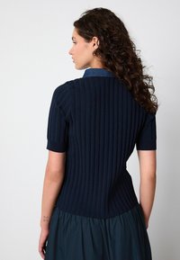 Mörkblå ribbad stickad topp med korta ärmar och en denimkrage. Har en figursydd silhuett och texturerad tyg. Bakvy visas.