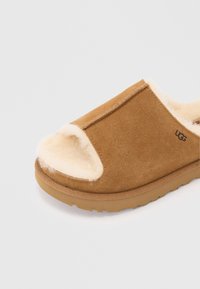 Chausson ouvert en daim marron avec doublure peluche crème et semelle en caoutchouc, logo UGG sur le côté.