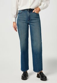 Vaqueros de mezclilla azul con cintura alta, pierna recta y costuras mínimas. Combinados con zuecos negros con tachuelas plateadas.